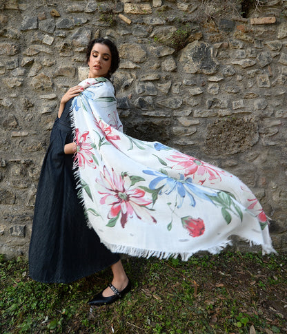Donna con abito scuro e sciarpa Foulard Bamboo De Nittis bianca, ampia con fiori rosa e blu dipinti, che svolazza al vento davanti a un muro di pietra grezza.


